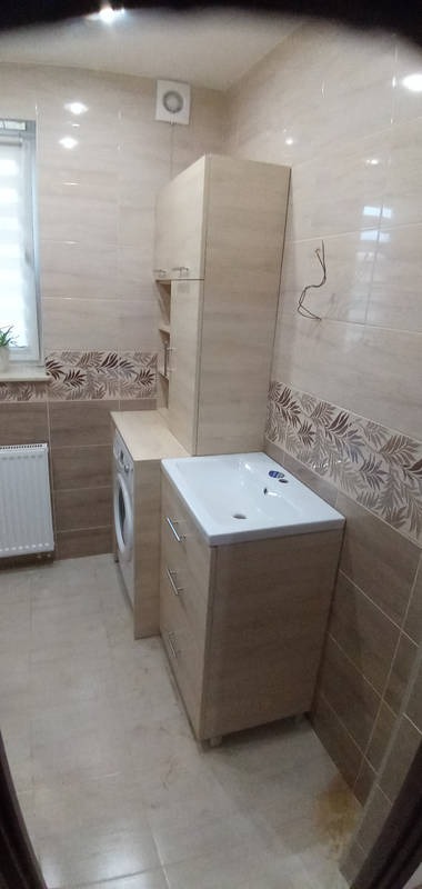 Jasna łazienka z umywalką w szafce z szufladami, pralką obok i szafką wiszącą. Beżowe płytki z motywem liści. Widoczny grzejnik i wentylator.