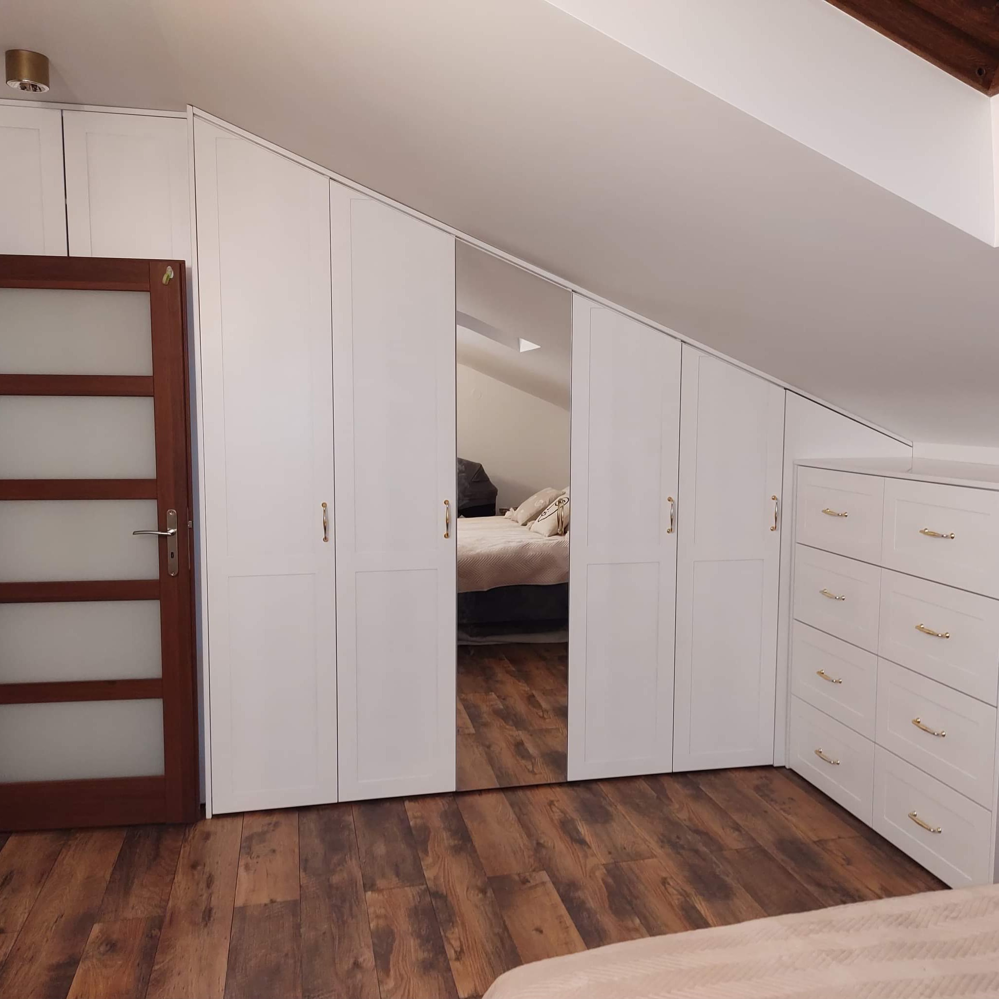 Biała szafa na wymiar ze złotymi uchwytami, wkomponowana w skos dachu na poddaszu. Lustrzane drzwi szafy odbijają wnętrze sypialni. Ciemna drewniana podłoga.
