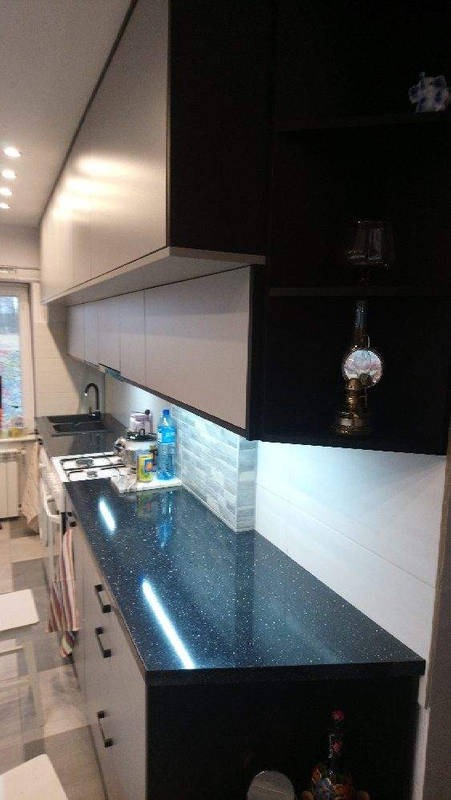 Nowoczesna kuchnia z czarnym blatem w gwiazdki i białymi szafkami. Oświetlenie LED podkreśla fakturę ściany i blatu. Na półkach dekoracje.