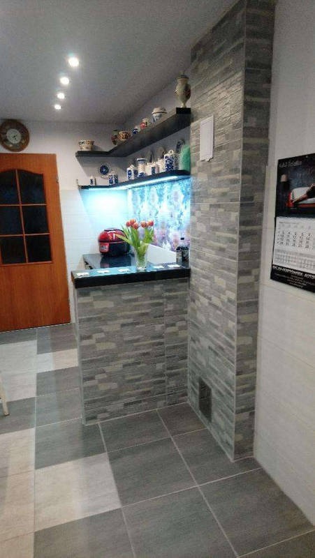 Wykończona kuchnia z barkiem, płytki imitujące kamień na ścianach i barku, półka z dekoracjami. Uwagę przykuwa akwarium wkomponowane w przestrzeń kuchenną.