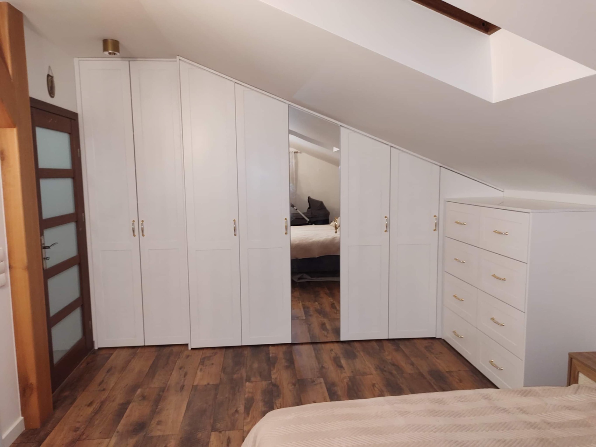 Biała szafa na wymiar ze złotymi uchwytami, wkomponowana w skos dachu na poddaszu, z lustrzanym frontem i komodą, na drewnianej podłodze.