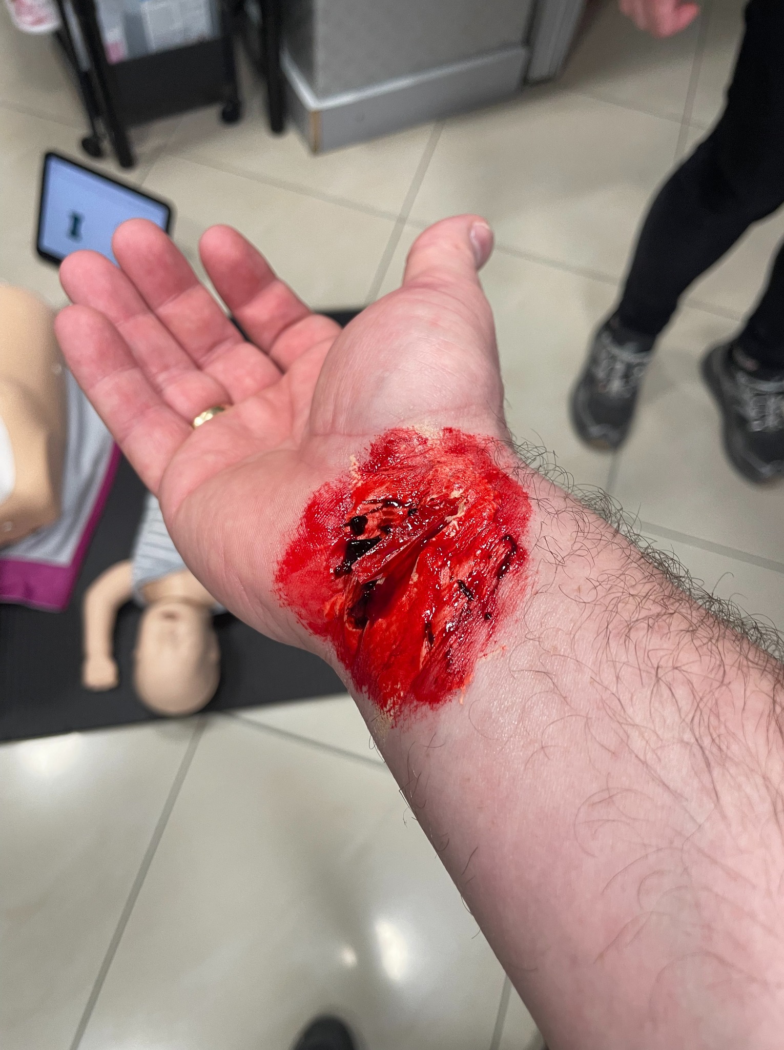 Realistyczna symulacja rany na przedramieniu, używana podczas szkoleń z pierwszej pomocy. W tle manekin, tablet i fragmenty sali szkoleniowej.