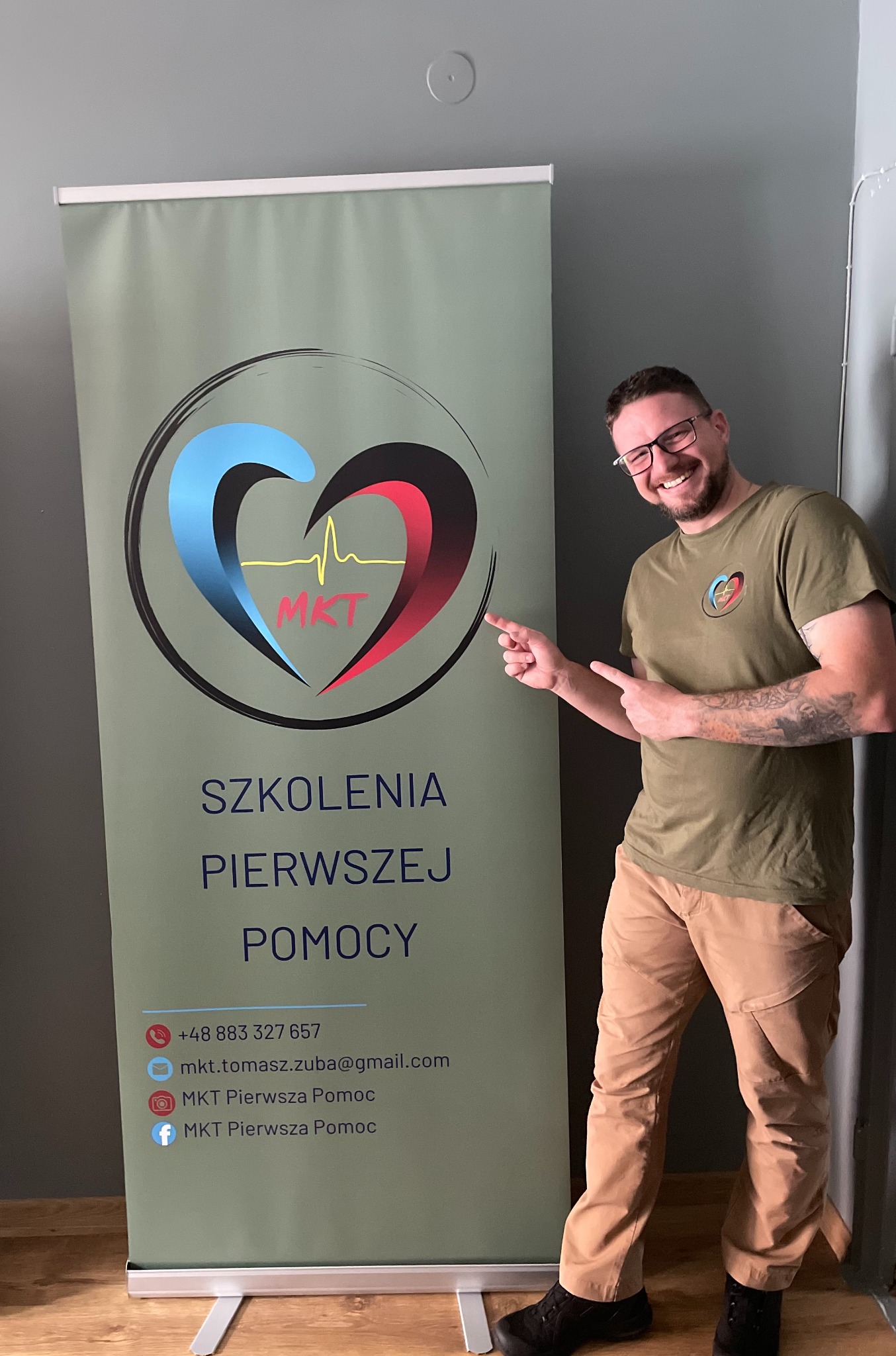 Uśmiechnięty mężczyzna w okularach wskazuje na roll-up z logo i napisem 'Szkolenia Pierwszej Pomocy'. Mężczyzna ma koszulkę z logo firmy. Widoczne dane kontaktowe.