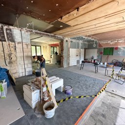 REMONTUJ Andrzej Tomanek - Wnętrze w trakcie remontu: pracownik miesza zaprawę, widoczne cegły, bloczki, narzędzia, zabezpieczenia podłogi folią i taśmą ostrzegawczą. Prace wykończeniowe w toku.