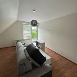 REMONTUJ Andrzej Tomanek - Jasne poddasze z drewnianą podłogą, szarą sofą i białymi ścianami. Okno z żaluzjami wpuszcza światło. Nowoczesna lampa sufitowa i biała szafka RTV uzupełniają minimalistyczny wystrój.