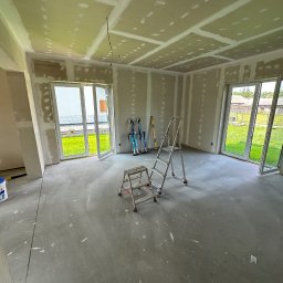 REMONTUJ Andrzej Tomanek - Wnętrze w trakcie remontu: ściany i sufit z płyt karton-gips, widoczne łączenia szpachlowane, drabina, szczudła malarskie. Dwa okna balkonowe z widokiem na zieleń.
