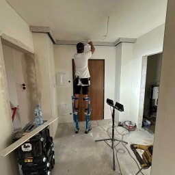 REMONTUJ Andrzej Tomanek - Praca na wysokościach: mężczyzna na szczudłach montuje konstrukcję sufitu podwieszanego w jasnym wnętrzu. Widoczne narzędzia, oświetlenie i fragmenty ścian w trakcie wykończenia.