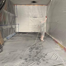 REMONTUJ Andrzej Tomanek - Pracownik w kombinezonie ochronnym maluje sufit i ściany pomieszczenia za pomocą pistoletu natryskowego. Podłoga i ściany zabezpieczone folią ochronną.