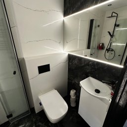 REMONTUJ Andrzej Tomanek - Nowoczesna łazienka z czarno-białymi płytkami imitującymi marmur, podświetlanym lustrem i minimalistyczną armaturą. Widoczna kabina prysznicowa i toaleta.