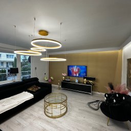 REMONTUJ Andrzej Tomanek - Nowoczesny salon z czarną sofą, złotym stolikiem kawowym i okrągłymi lampami wiszącymi. Ściana akcentowa w kolorze złota. Widok na zewnątrz przez okno.
