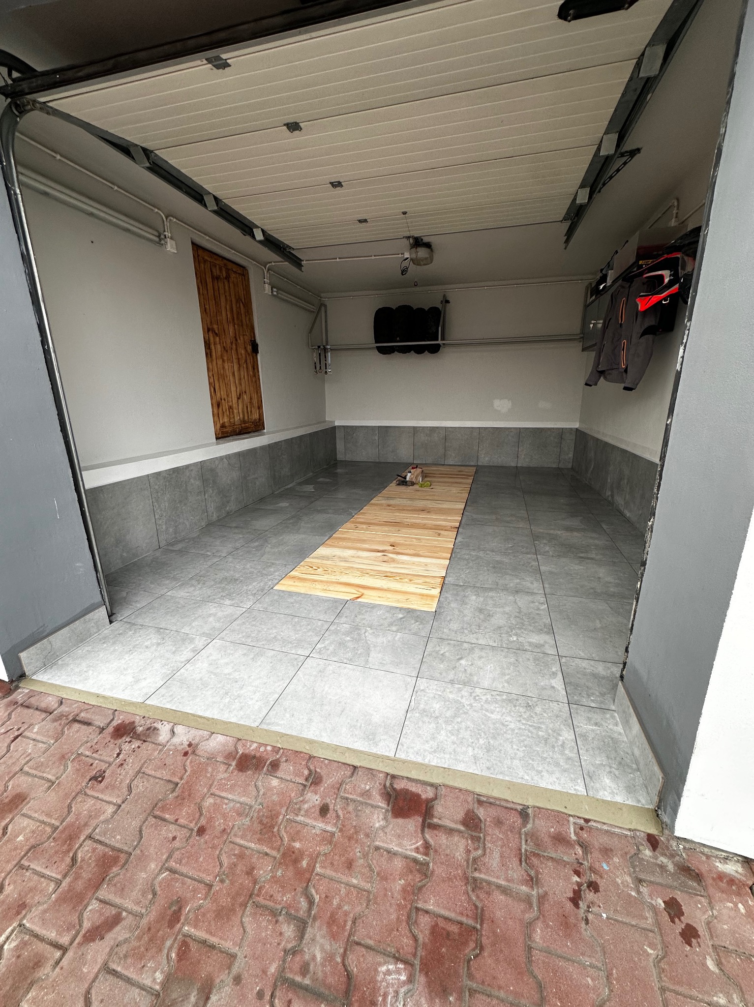 Wykończone wnętrze garażu z szarymi płytkami podłogowymi i drewnianą platformą na środku. Widoczne białe ściany, drewniane drzwi i elementy wyposażenia.