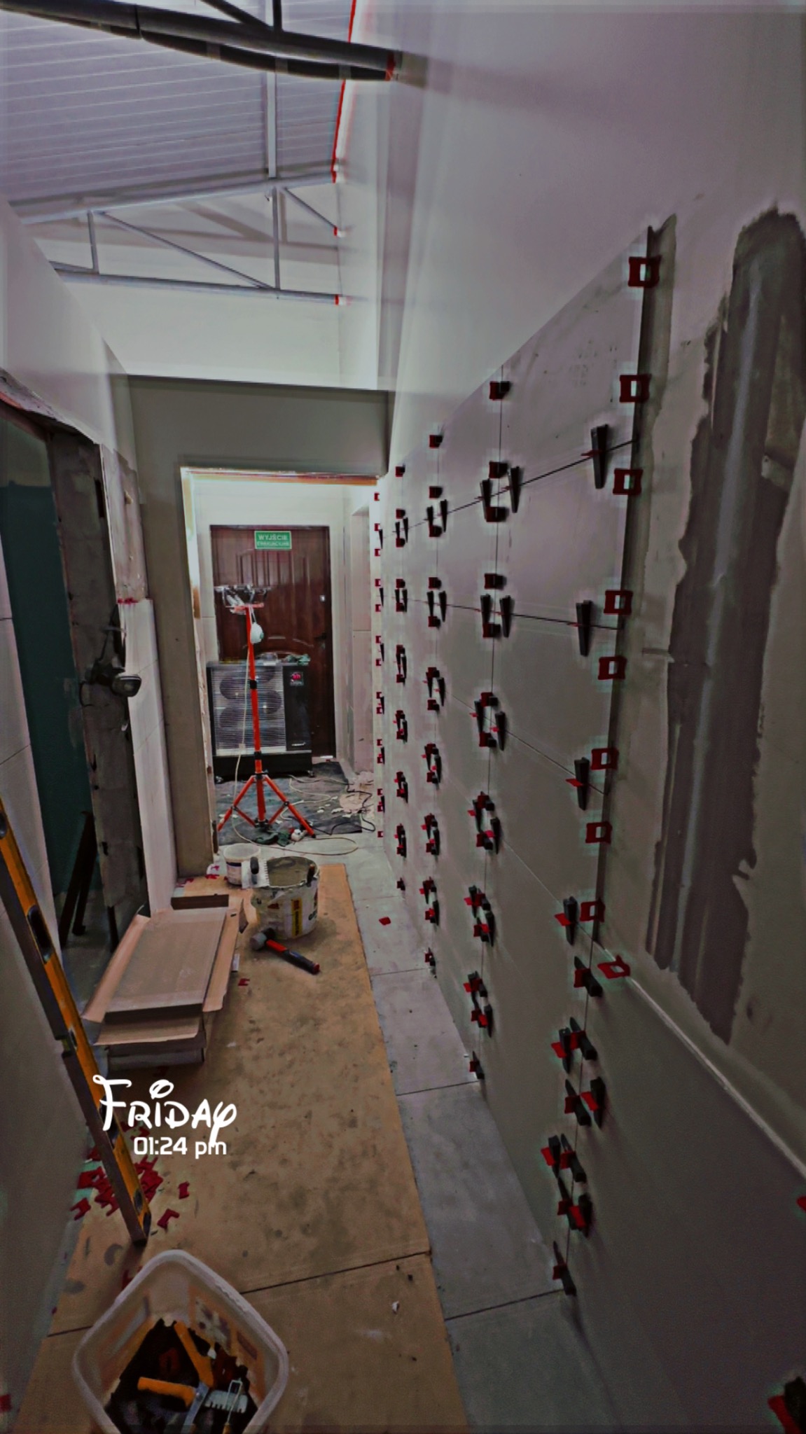 Układanie płytek na ścianie z użyciem systemu poziomowania. Widoczne narzędzia, wiadro z klejem i poziomica. Prace glazurnicze w trakcie realizacji.