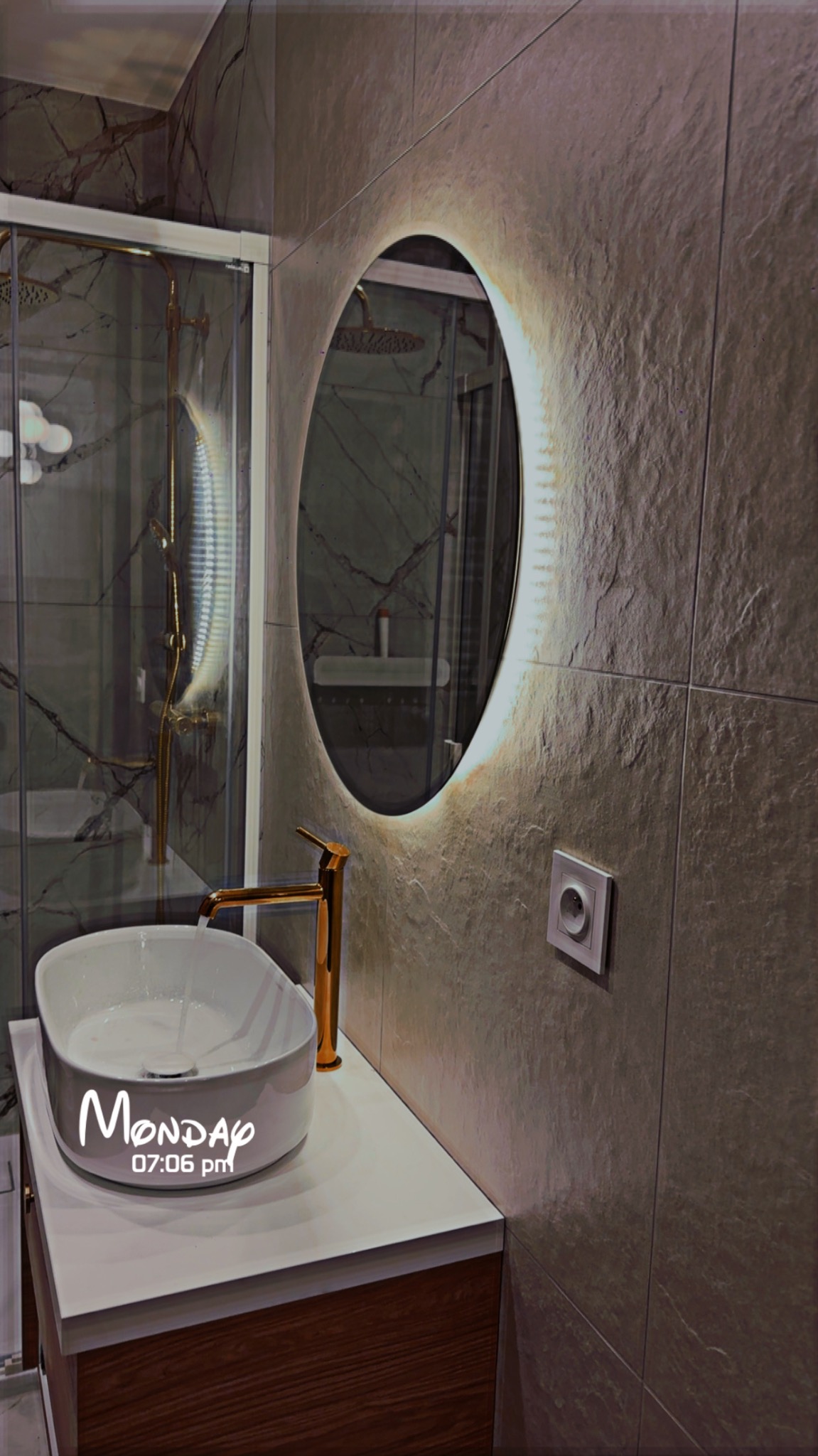 Nowoczesna łazienka z owalnym lustrem LED, białą umywalką nablatową i złotą armaturą. Widoczna kabina prysznicowa z marmurowym wykończeniem.