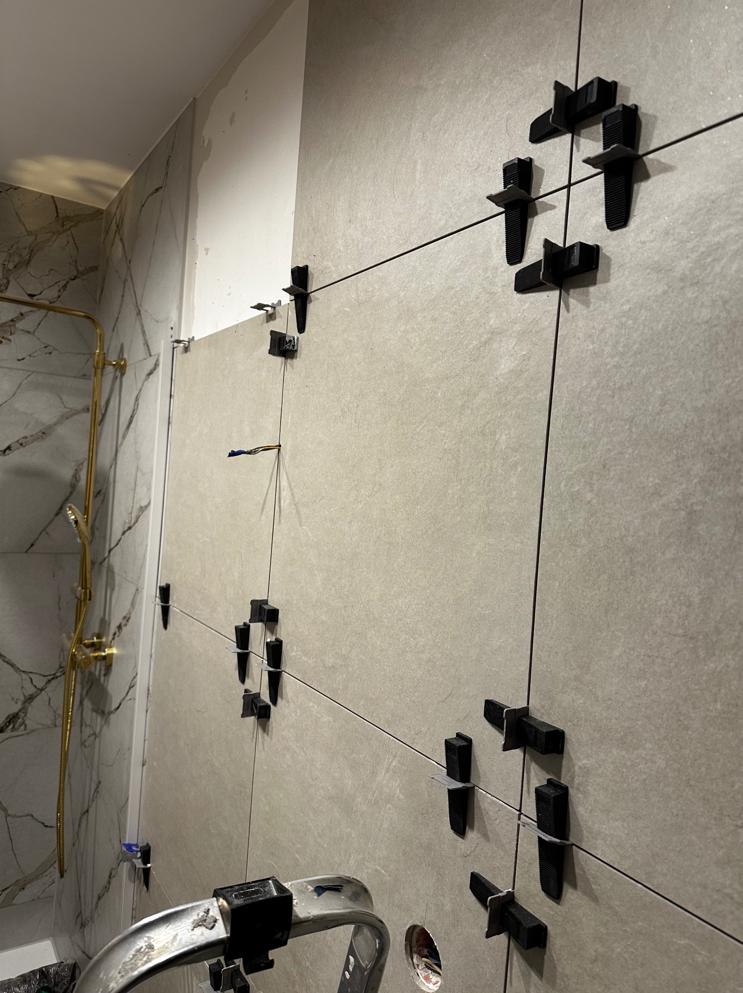 Układanie płytek na ścianie w łazience w Starachowicach. Widoczne krzyżyki dystansowe i system poziomowania płytek. Częściowo wykończona ściana z szarych płytek.