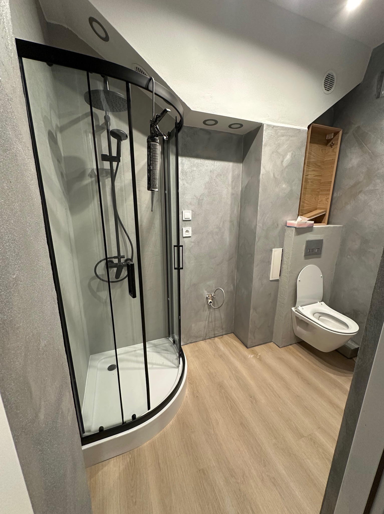 Nowoczesna łazienka po remoncie: kabina prysznicowa z czarnymi elementami, toaleta podwieszana i ściany wykończone w szarościach. Widoczna puszka z pianką montażową na kabinie.