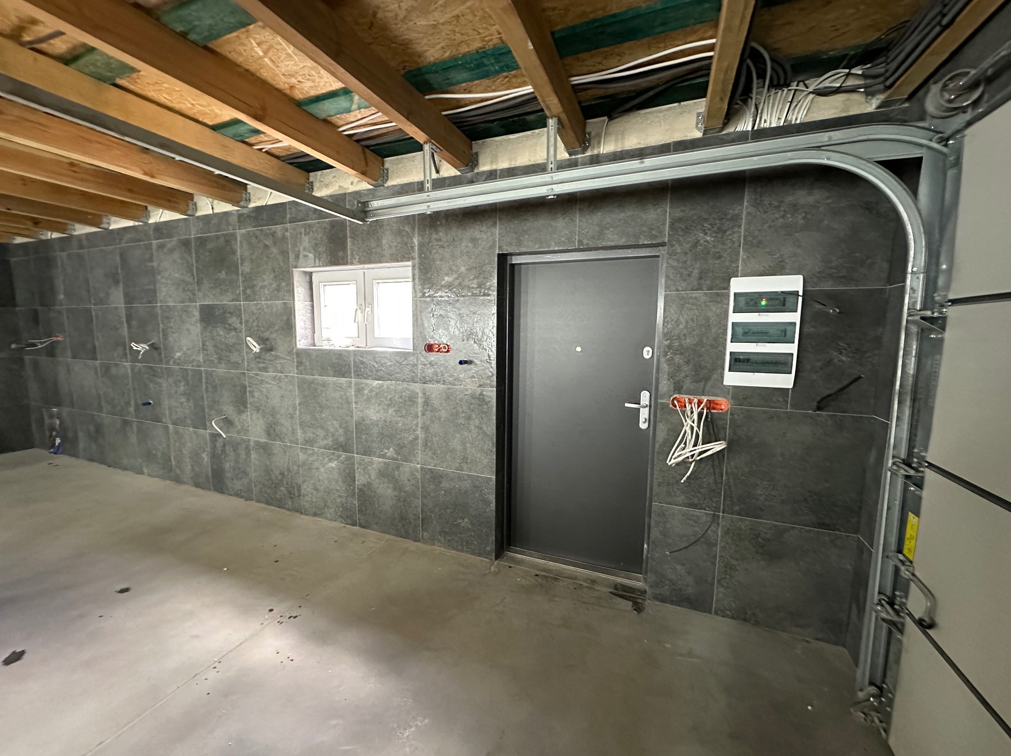 Wykończone wnętrze garażu z szarymi płytkami na ścianach, widoczną instalacją elektryczną i metalową bramą garażową. Okno i drzwi wejściowe w tle.