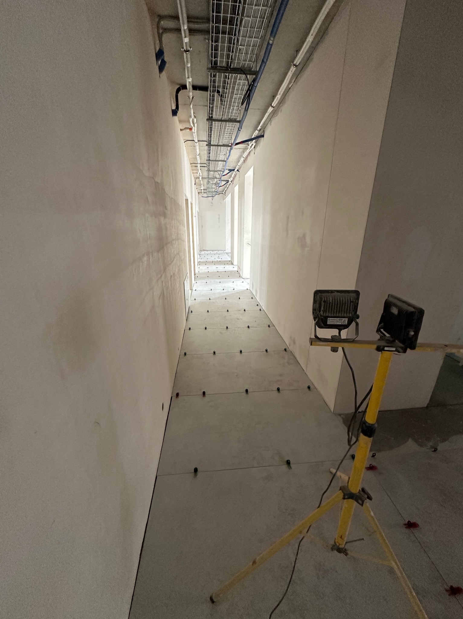 Korytarz w trakcie układania płytek podłogowych, widoczne krzyżyki dystansowe i lampa budowlana. Ściany przygotowane do malowania lub szpachlowania.