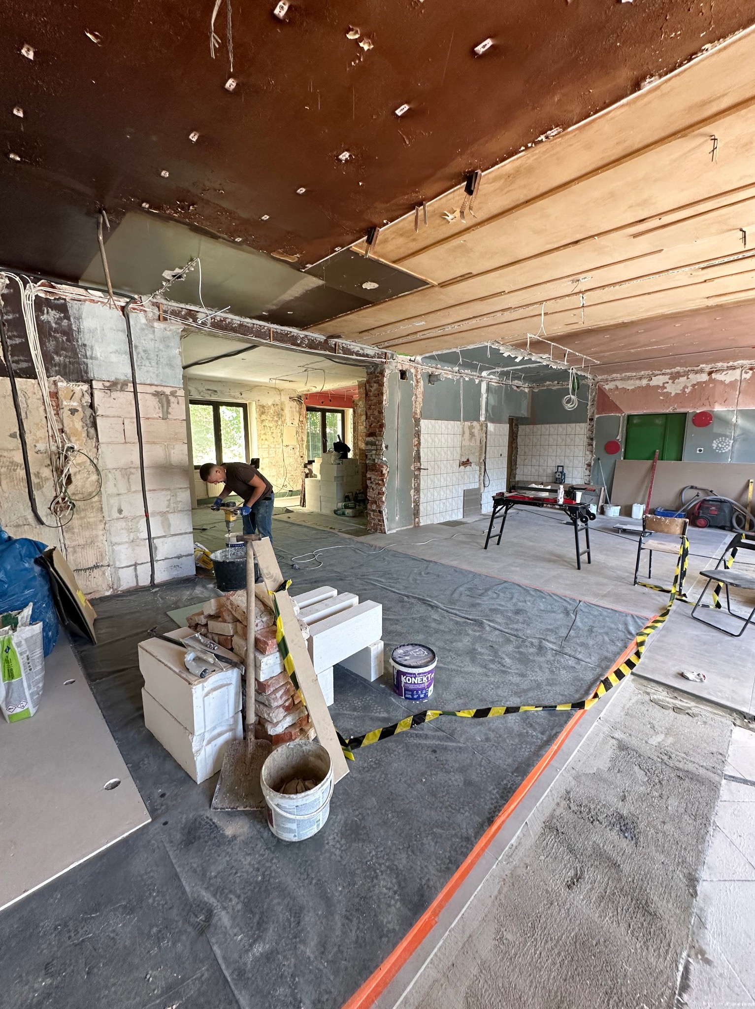 Wnętrze w trakcie remontu: pracownik miesza zaprawę, widoczne cegły, bloczki, narzędzia, zabezpieczenia podłogi folią i taśmą ostrzegawczą. Prace wykończeniowe w toku.