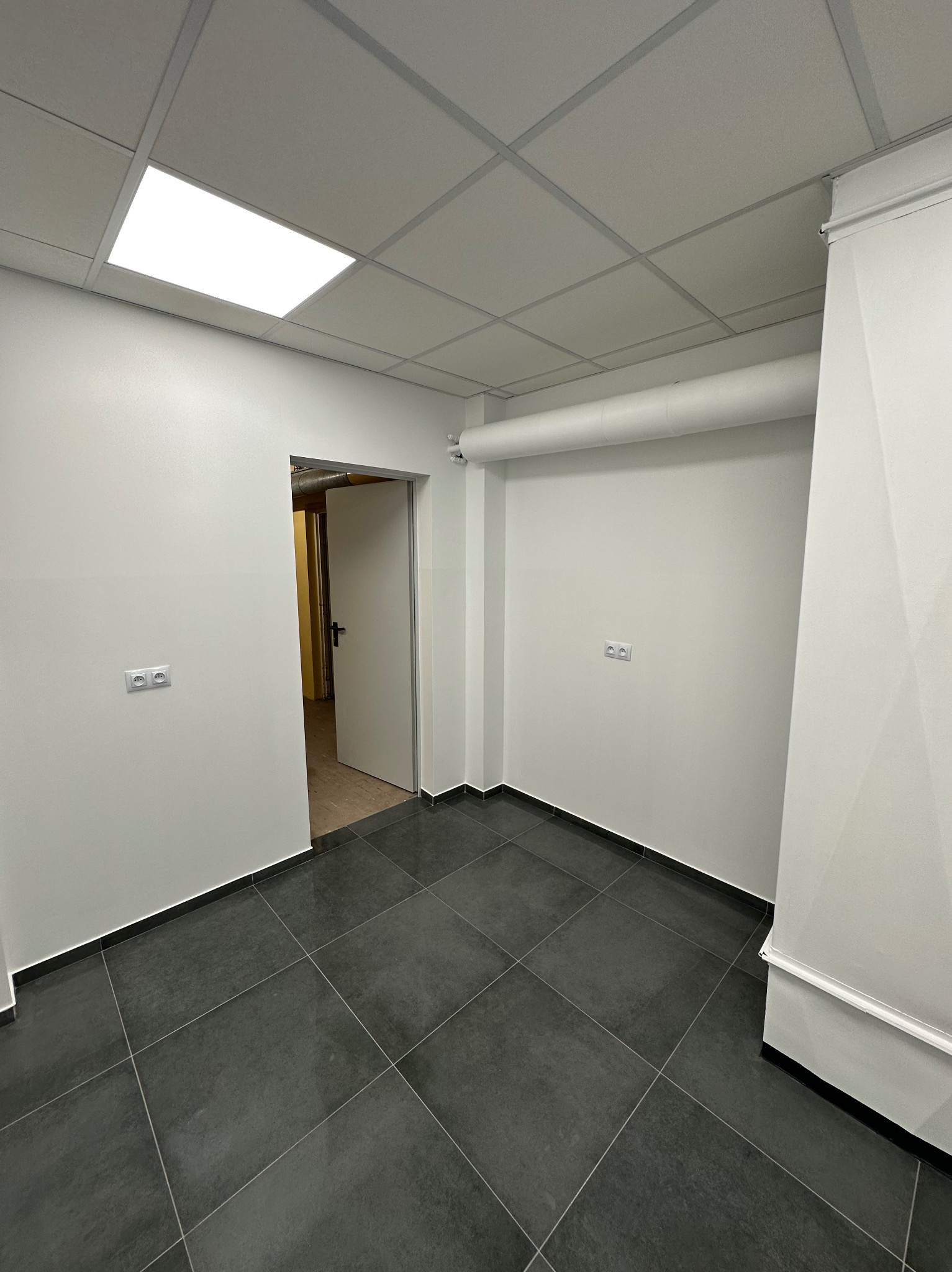 Wnętrze biura po remoncie: białe ściany, ciemna podłoga z płytek, sufit podwieszany z oświetleniem. Widoczne gniazdka elektryczne i otwarte drzwi do innego pomieszczenia.