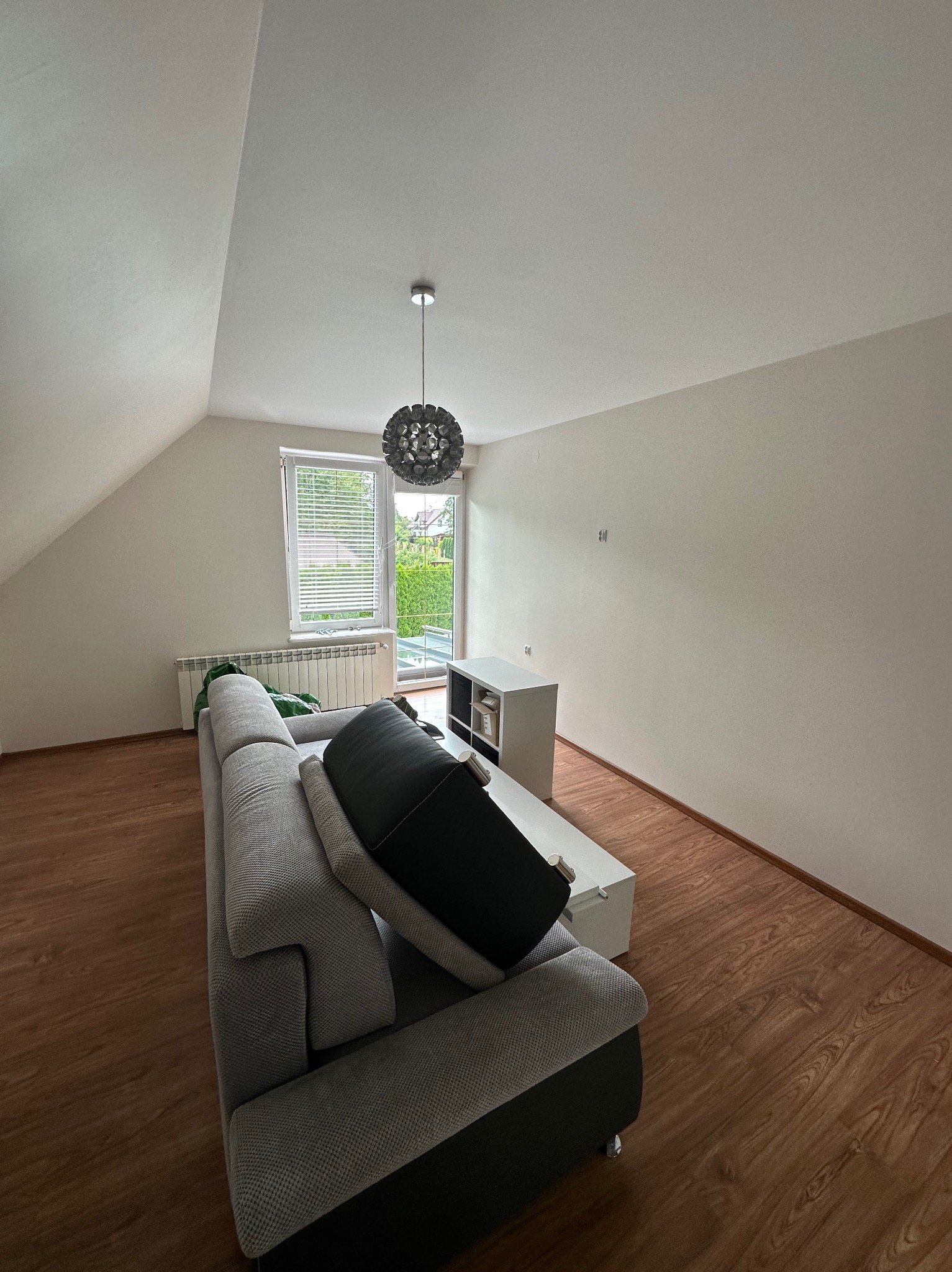 Jasne poddasze z drewnianą podłogą, szarą sofą i białymi ścianami. Okno z żaluzjami wpuszcza światło. Nowoczesna lampa sufitowa i biała szafka RTV uzupełniają minimalistyczny wystrój.