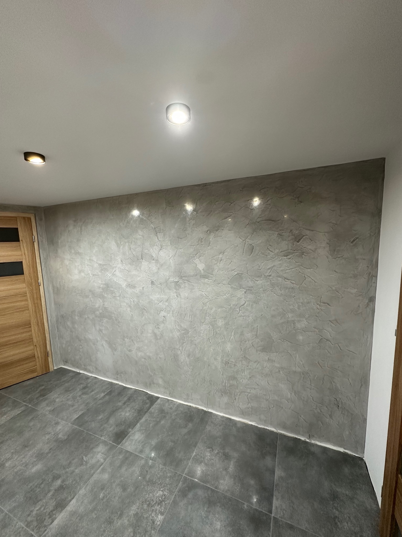 Szara ściana z efektem dekoracyjnym w pokoju z szarą podłogą z płytek. Widoczne drzwi z drewnianą okleiną i czarnymi wstawkami oraz oświetlenie sufitowe.