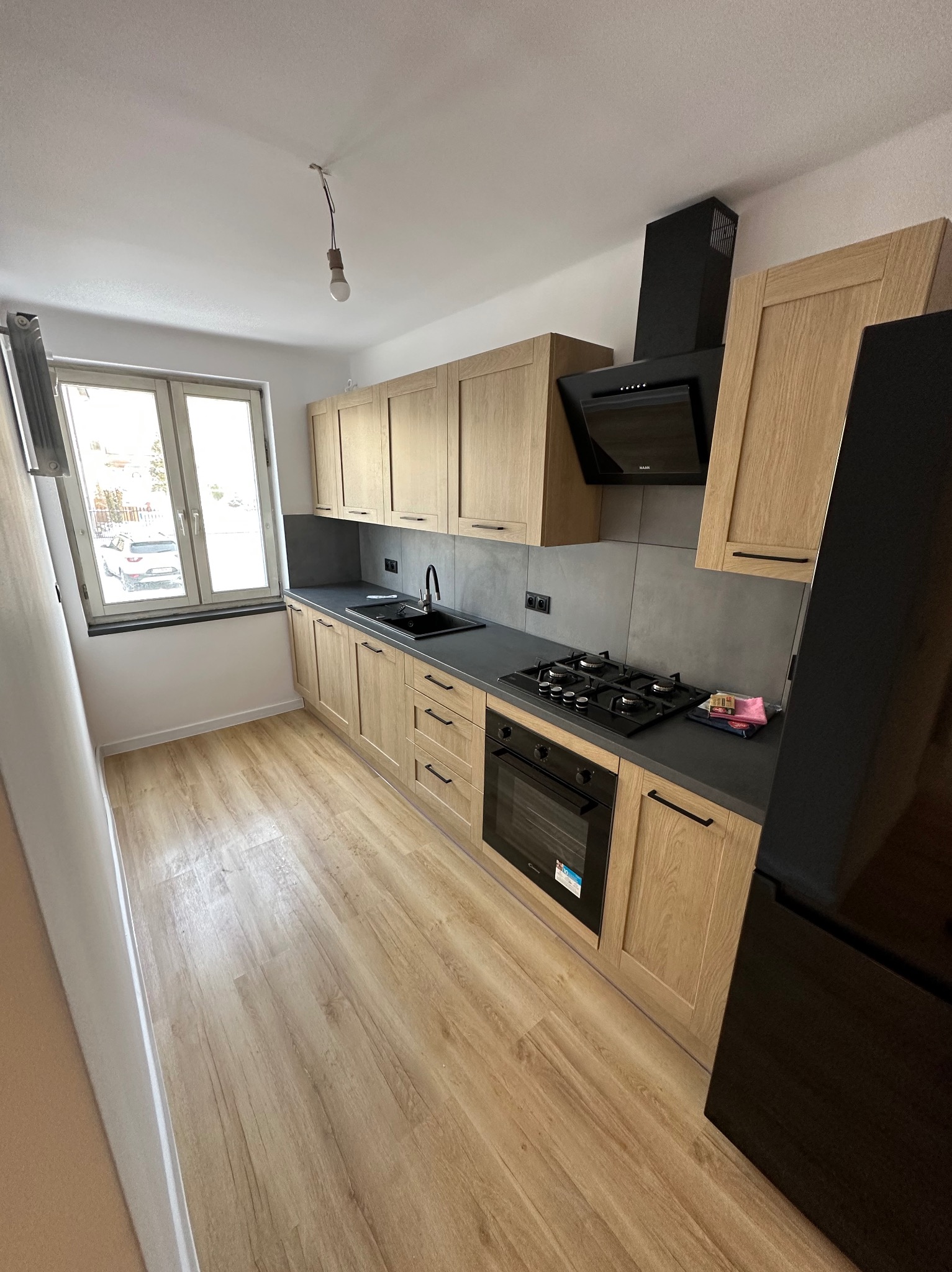 Jasna kuchnia z drewnianymi szafkami, czarnym blatem i sprzętem AGD. Widoczne okno z parapetem i podłoga imitująca drewno. Minimalistyczny design z wiszącą żarówką.