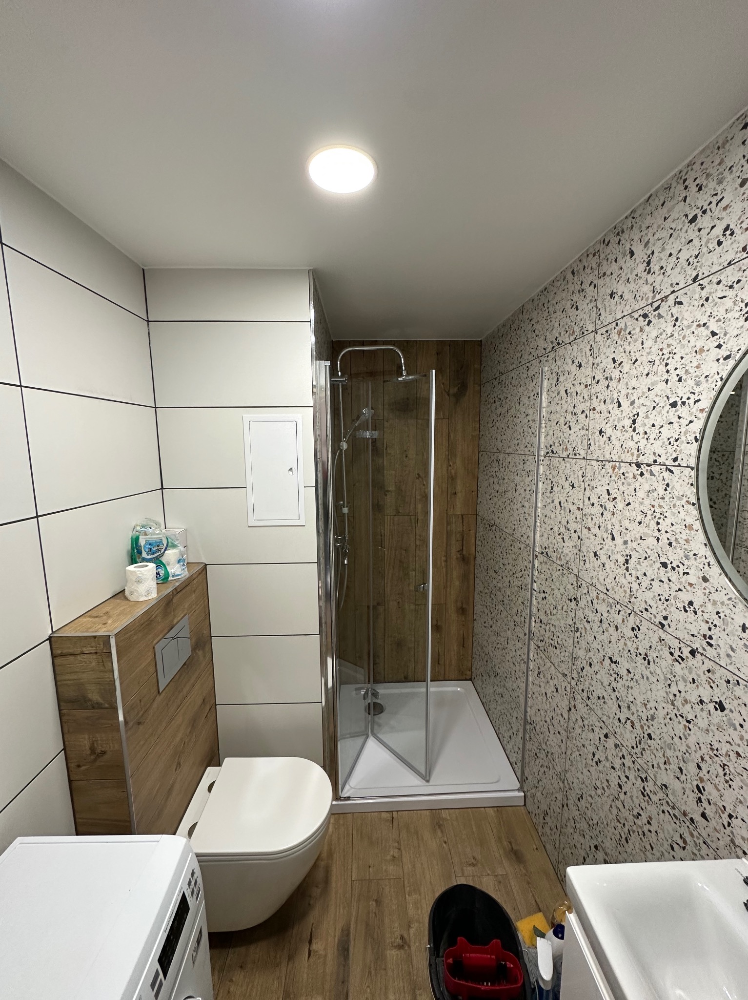 Wykończona łazienka z kabiną prysznicową, toaletą podwieszaną i umywalką. Ściany wyłożone płytkami w dwóch wzorach: białym z czarnymi fugami i mozaikowym.