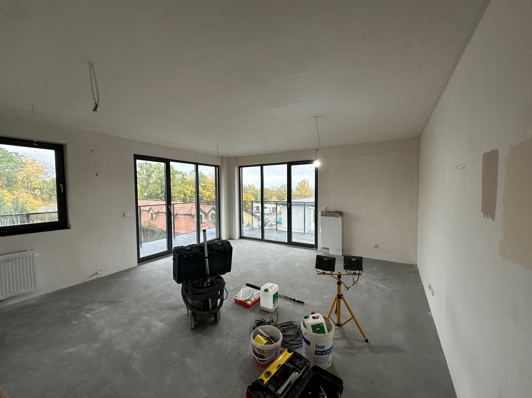 Wnętrze w trakcie szpachlowania: gładkie ściany, duże okna balkonowe, narzędzia malarskie, lampa na statywie. Widoczne ślady po łataniu ścian.