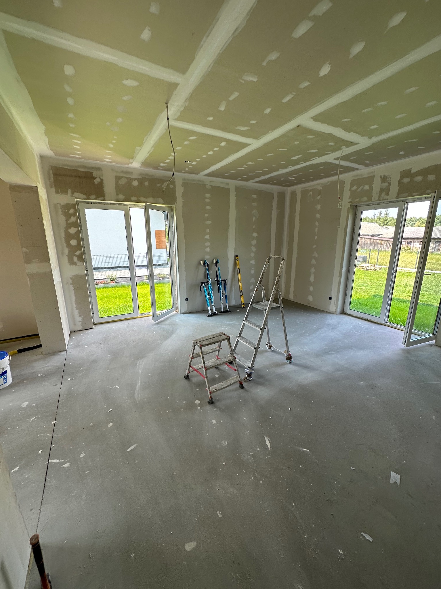 Wnętrze w trakcie remontu: ściany i sufit z płyt karton-gips, widoczne łączenia szpachlowane, drabina, szczudła malarskie. Dwa okna balkonowe z widokiem na zieleń.