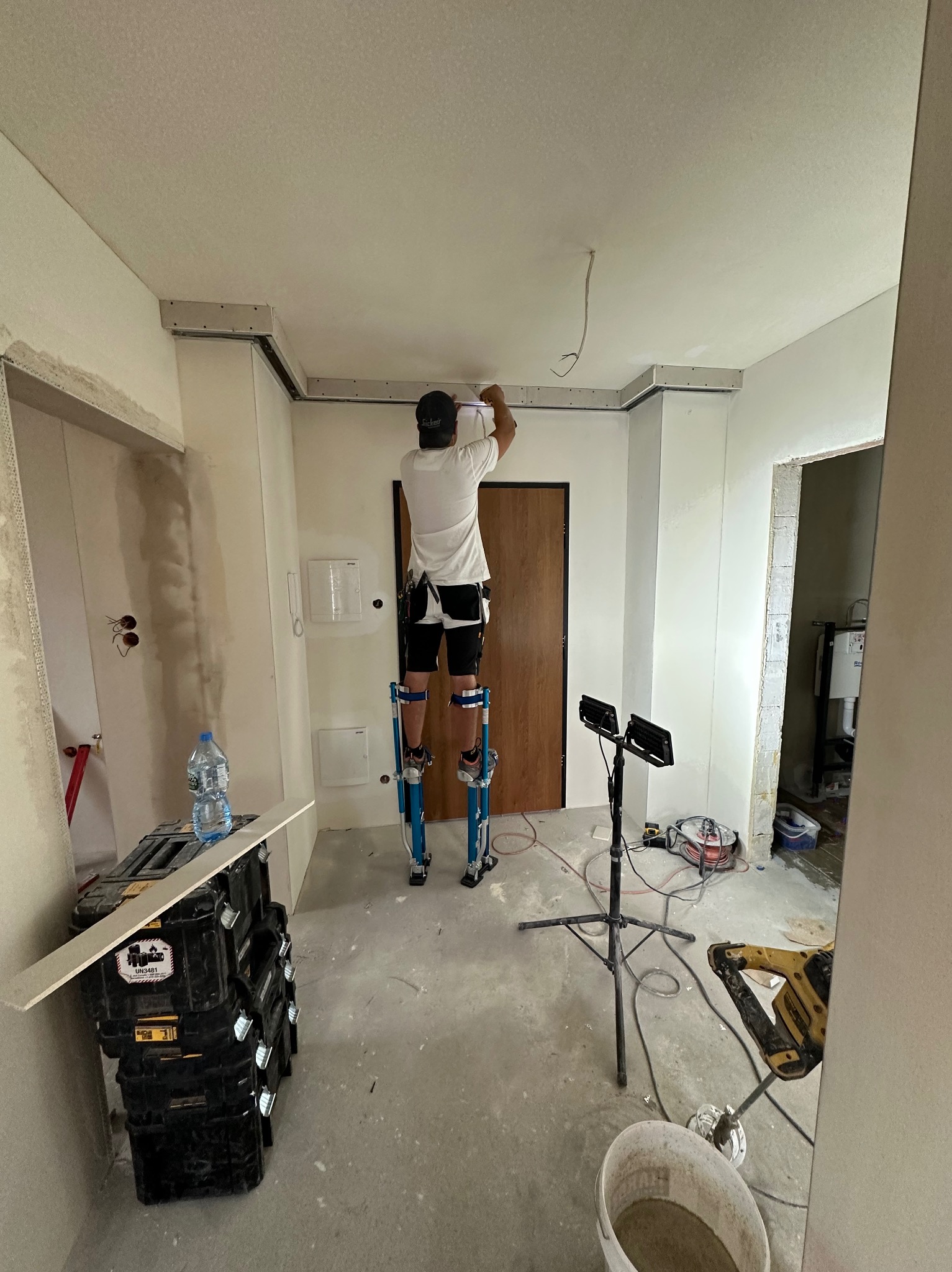 Praca na wysokościach: mężczyzna na szczudłach montuje konstrukcję sufitu podwieszanego w jasnym wnętrzu. Widoczne narzędzia, oświetlenie i fragmenty ścian w trakcie wykończenia.