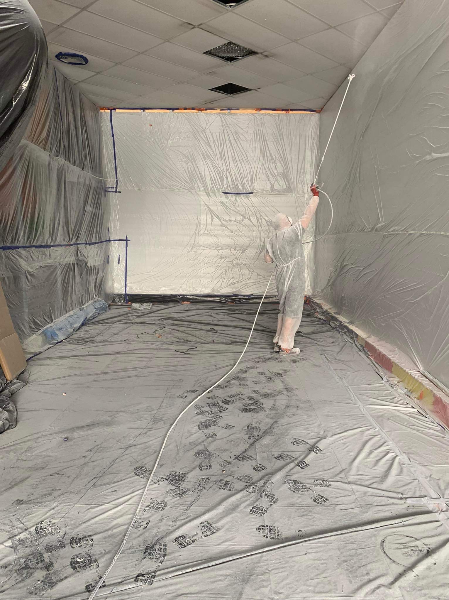 Pracownik w kombinezonie ochronnym maluje sufit i ściany pomieszczenia za pomocą pistoletu natryskowego. Podłoga i ściany zabezpieczone folią ochronną.