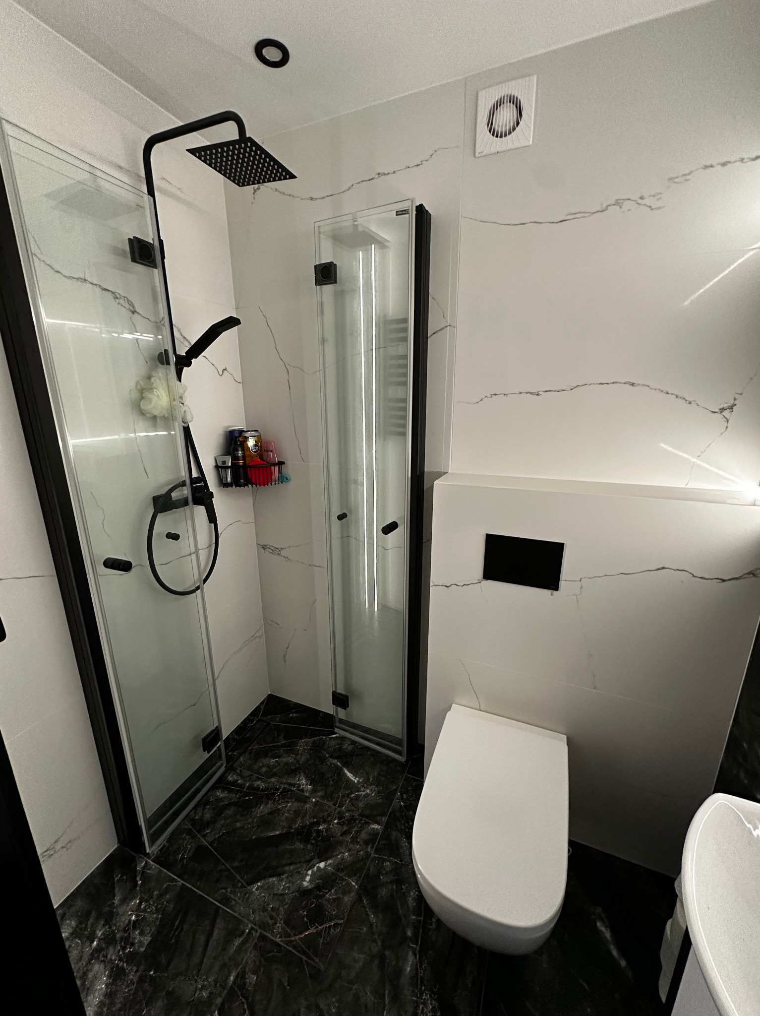 Nowoczesna łazienka z kabiną prysznicową typu walk-in, czarną armaturą i marmurową posadzką. Minimalistyczny design z akcentami czerni i bieli. Widoczny fragment toalety.