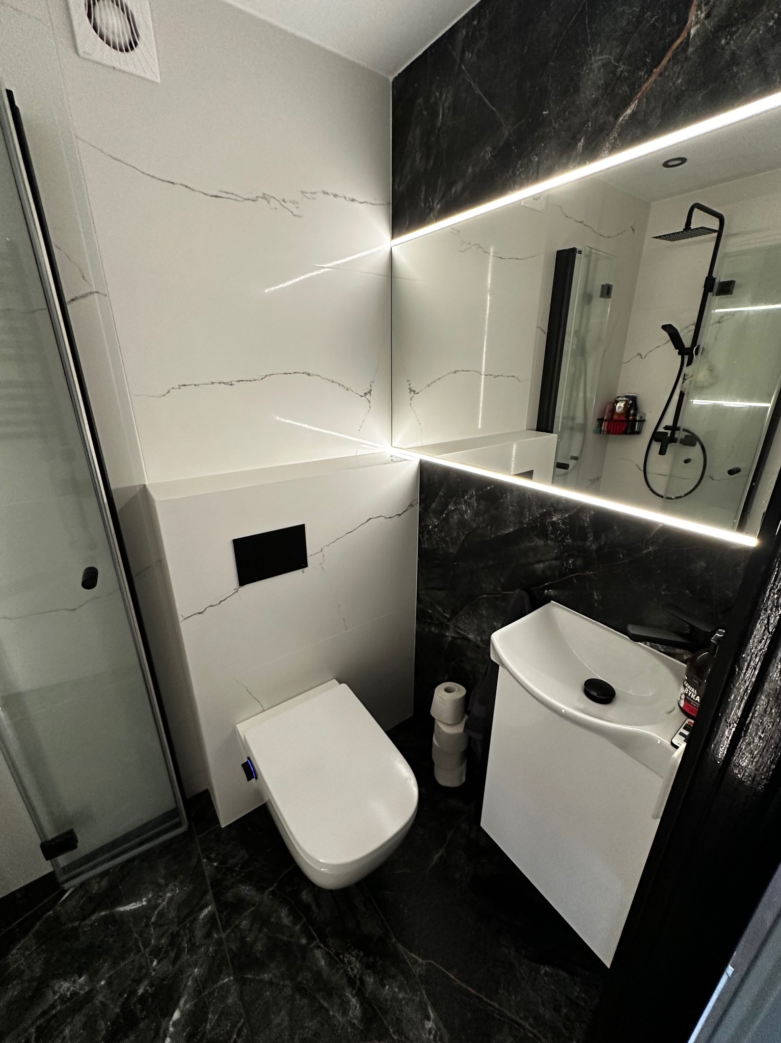 Nowoczesna łazienka z czarno-białymi płytkami imitującymi marmur, podświetlanym lustrem i minimalistyczną armaturą. Widoczna kabina prysznicowa i toaleta.