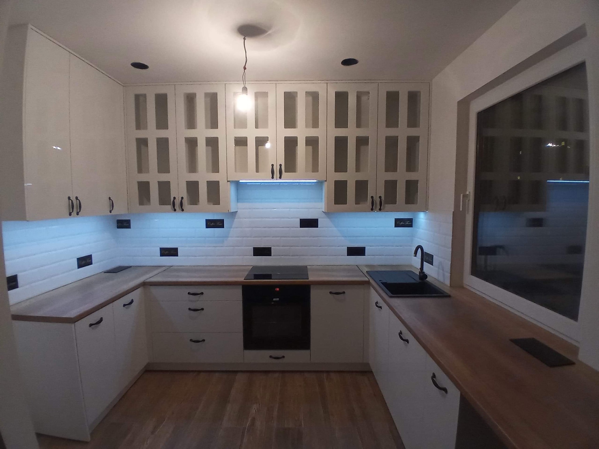 Jasna kuchnia w kształcie litery U z drewnianym blatem i białymi szafkami z przeszklonymi frontami. Nad blatem białe płytki z czarnymi akcentami.