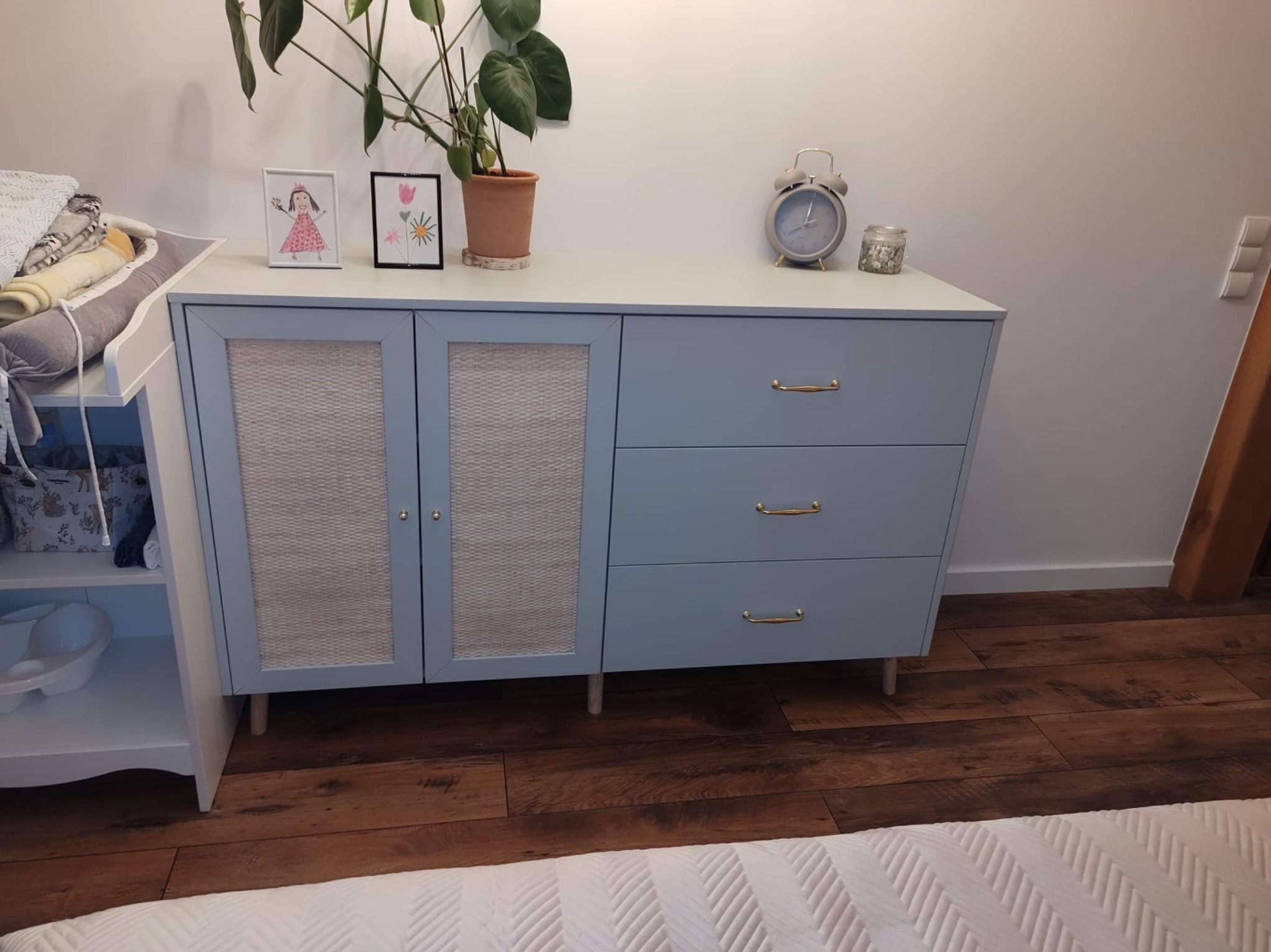 Niebieska komoda z trzema szufladami i dwoma drzwiczkami z rattanową wstawką, na której stoją dekoracje i roślina doniczkowa. Ustawiona na drewnianej podłodze.