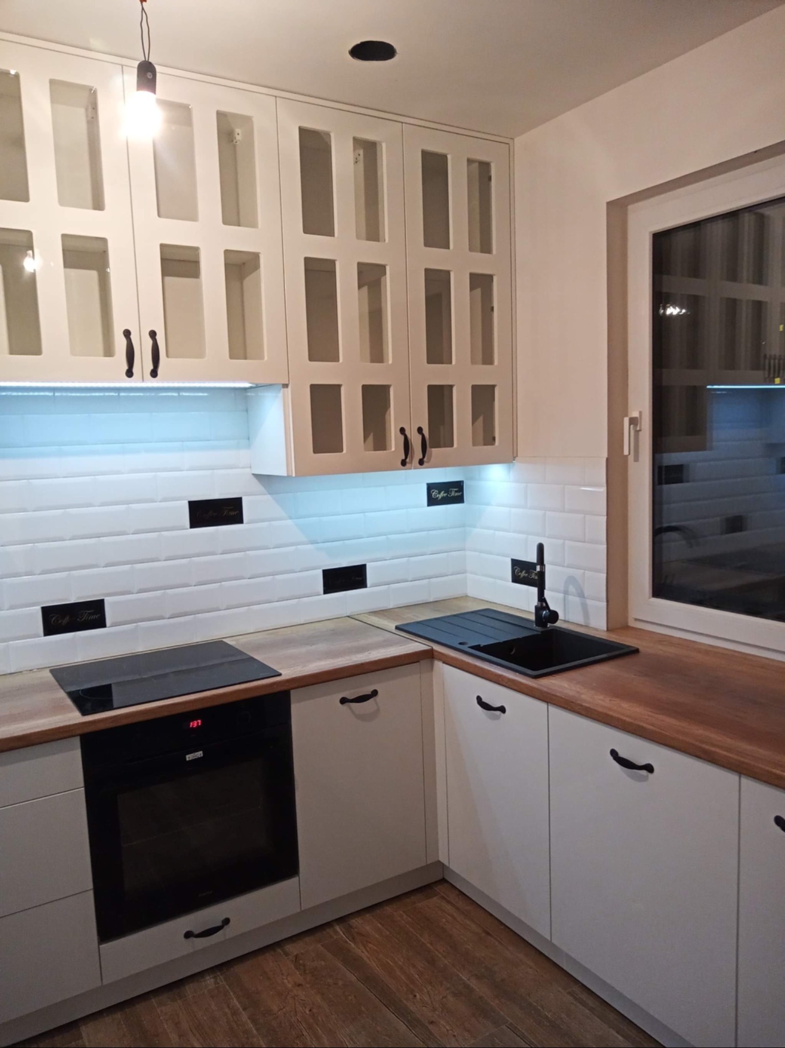 Jasna kuchnia z drewnianym blatem i białymi szafkami z przeszkleniami. Nad blatem białe płytki z czarnymi napisami. Nowoczesny design z oświetleniem LED.