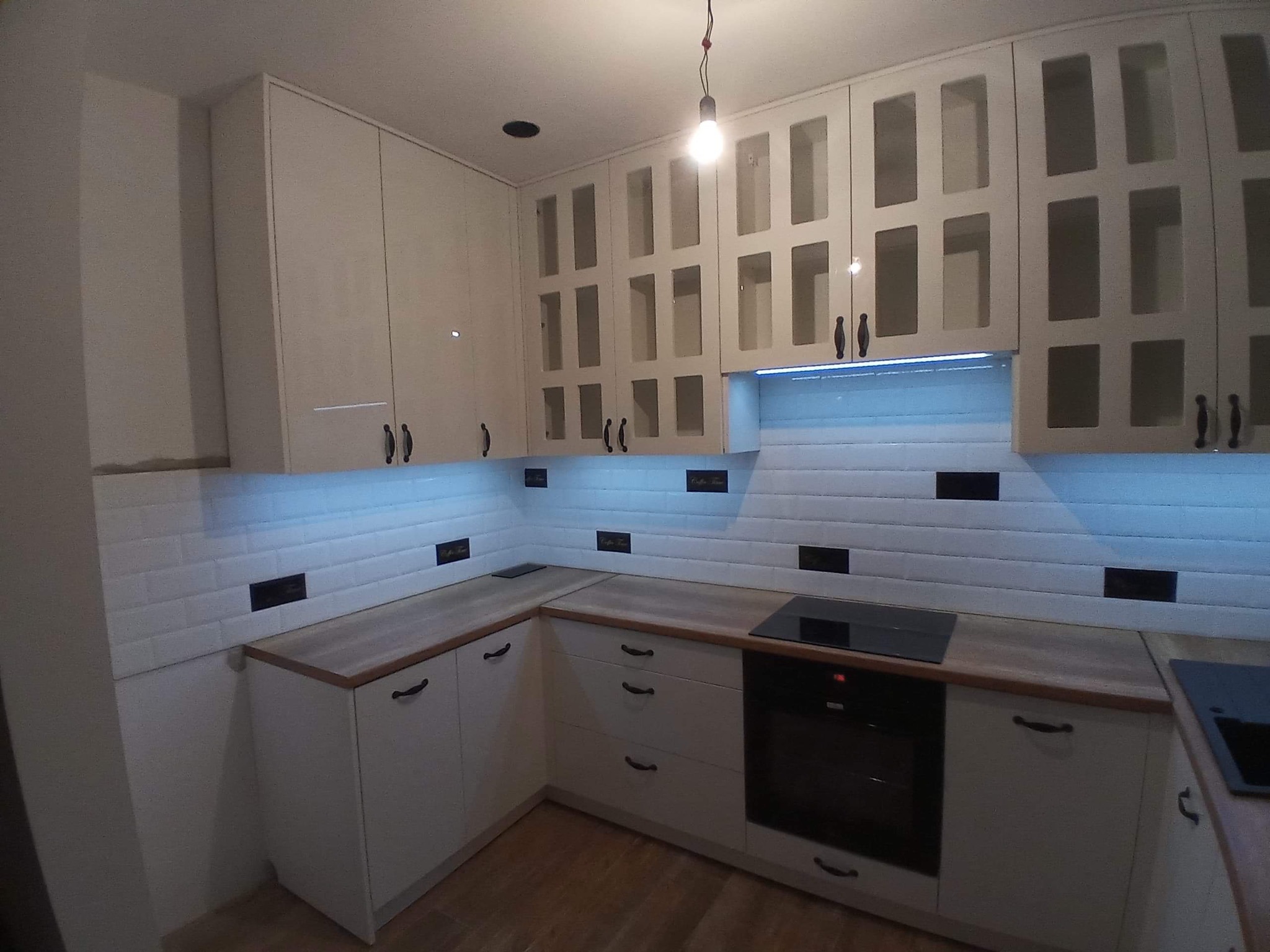 Biała kuchnia na wymiar z przeszklonymi szafkami wiszącymi i drewnianym blatem, oświetlona ledami, Radomsko. Widoczne gniazdka na ścianie z białej cegły.