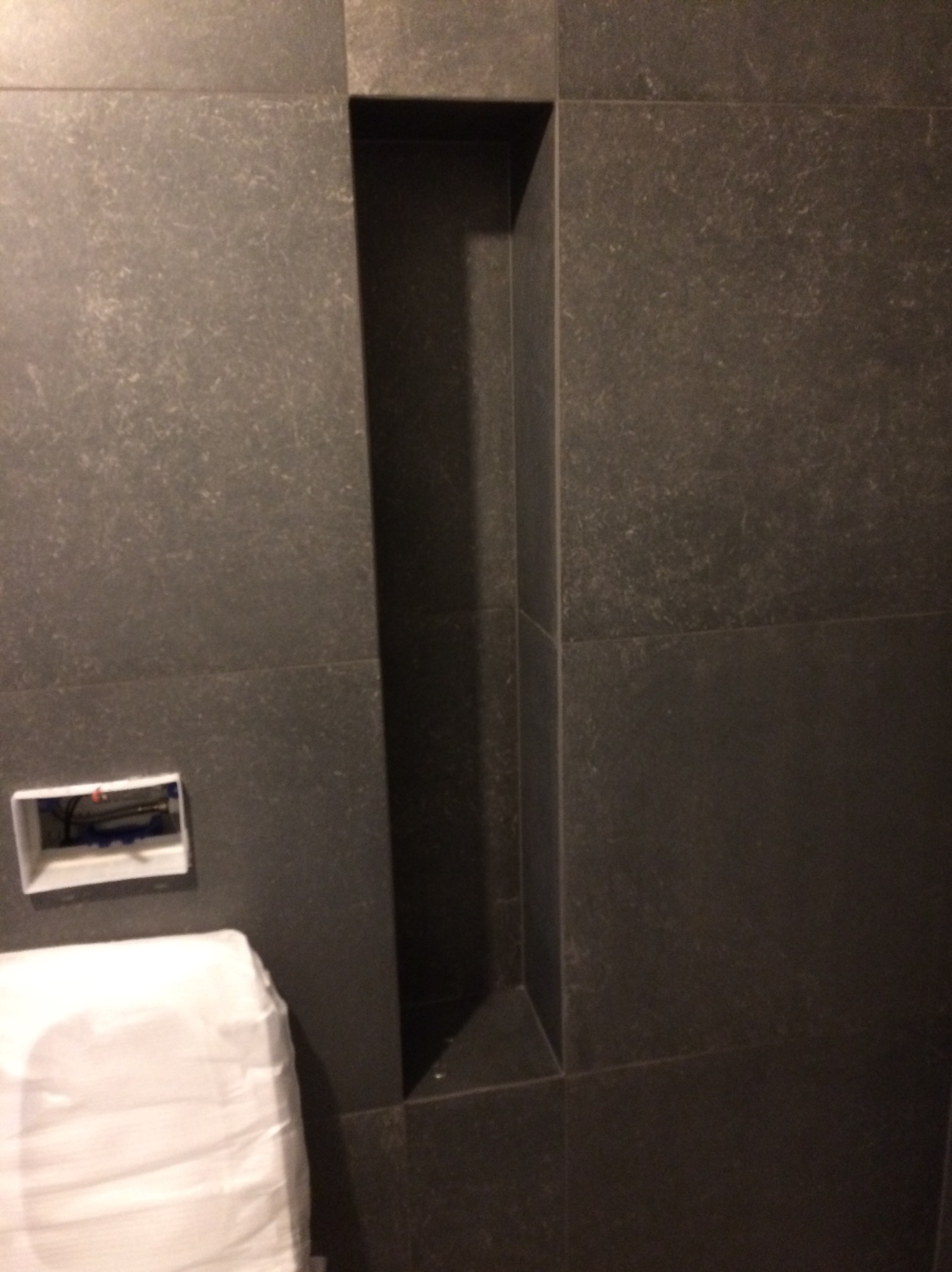 Ciemne, matowe płytki w łazience z wnęką i przygotowanym miejscem na toaletę. Widoczne kable elektryczne. Minimalistyczny design.