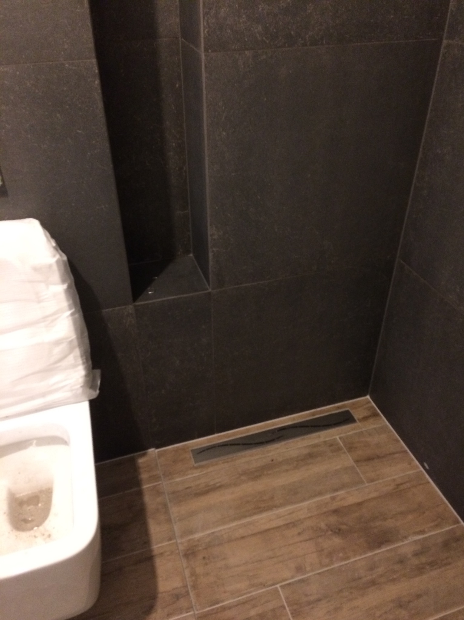 Nowoczesna łazienka z ciemnymi, matowymi płytkami i drewnopodobną podłogą. Uwagę przykuwa minimalistyczny odpływ liniowy i toaleta zabezpieczona folią ochronną.