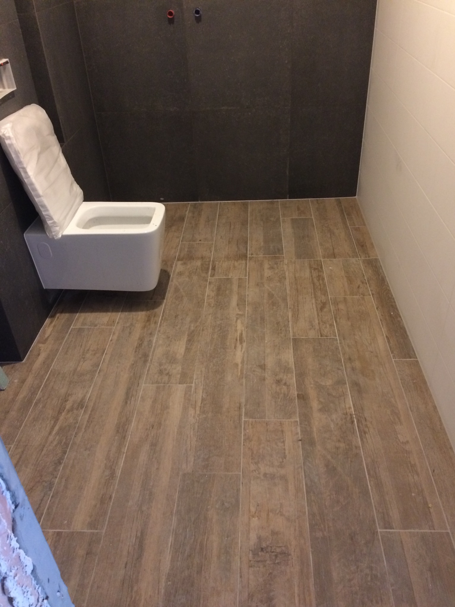 Nowoczesna łazienka z podwieszaną toaletą w folii ochronnej, brązowe płytki imitujące drewno na podłodze, ciemne płytki na ścianie i biała ściana z poziomymi panelami.