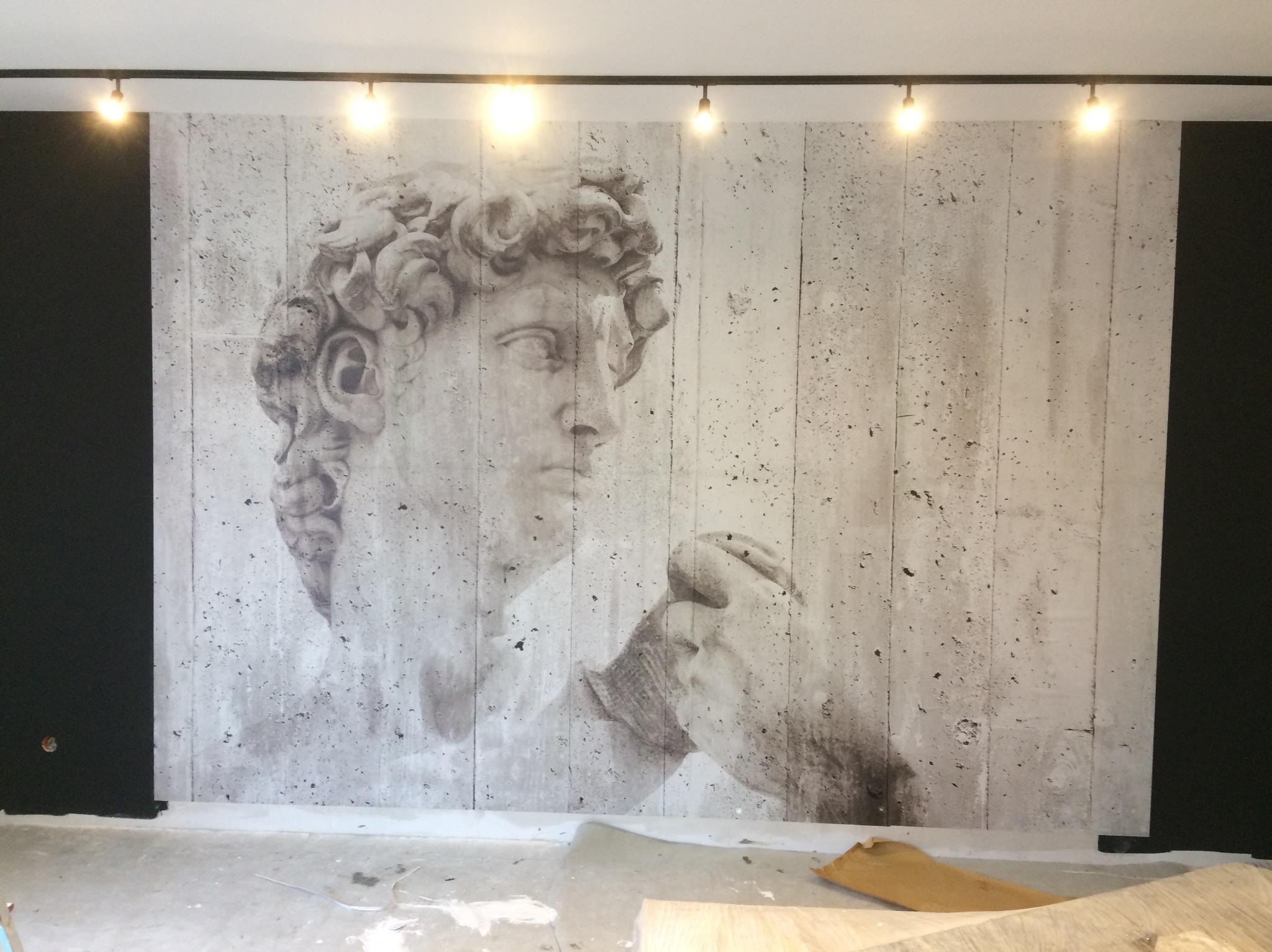 Ściana z fototapetą imitującą beton z wizerunkiem rzeźby Dawida, oświetlona punktowymi lampami sufitowymi. Widoczne fragmenty podłogi w trakcie prac wykończeniowych.