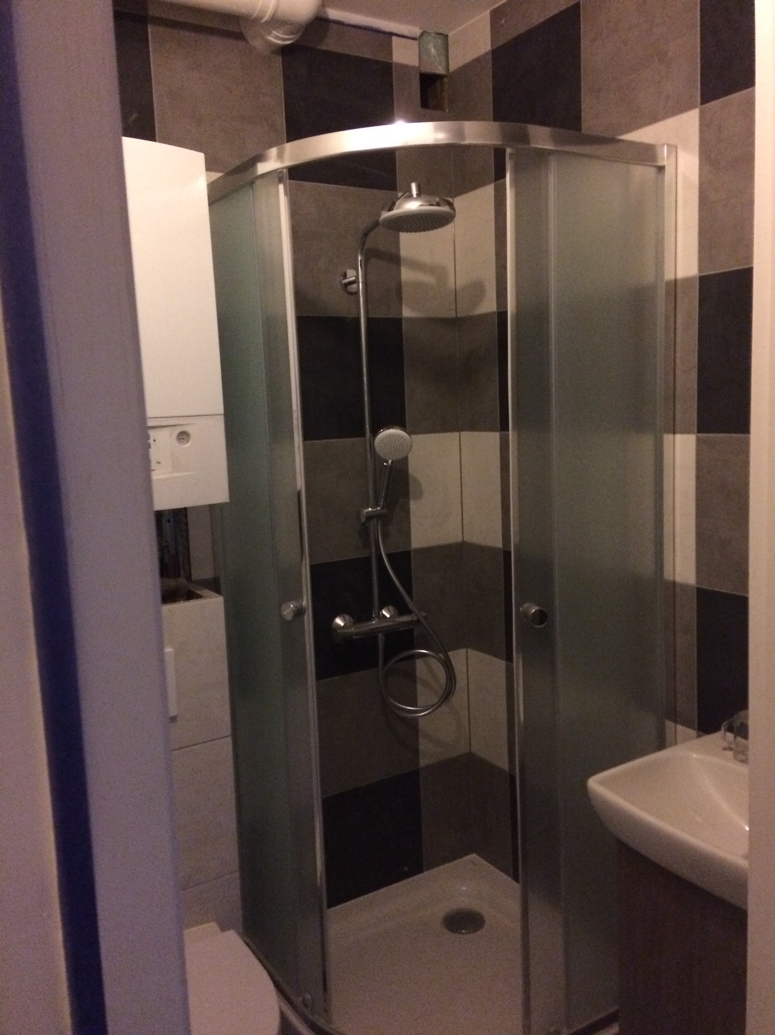 Nowoczesna kabina prysznicowa z szarymi płytkami w szachownicę. Widoczny bojler i umywalka. Niewielka łazienka po remoncie.