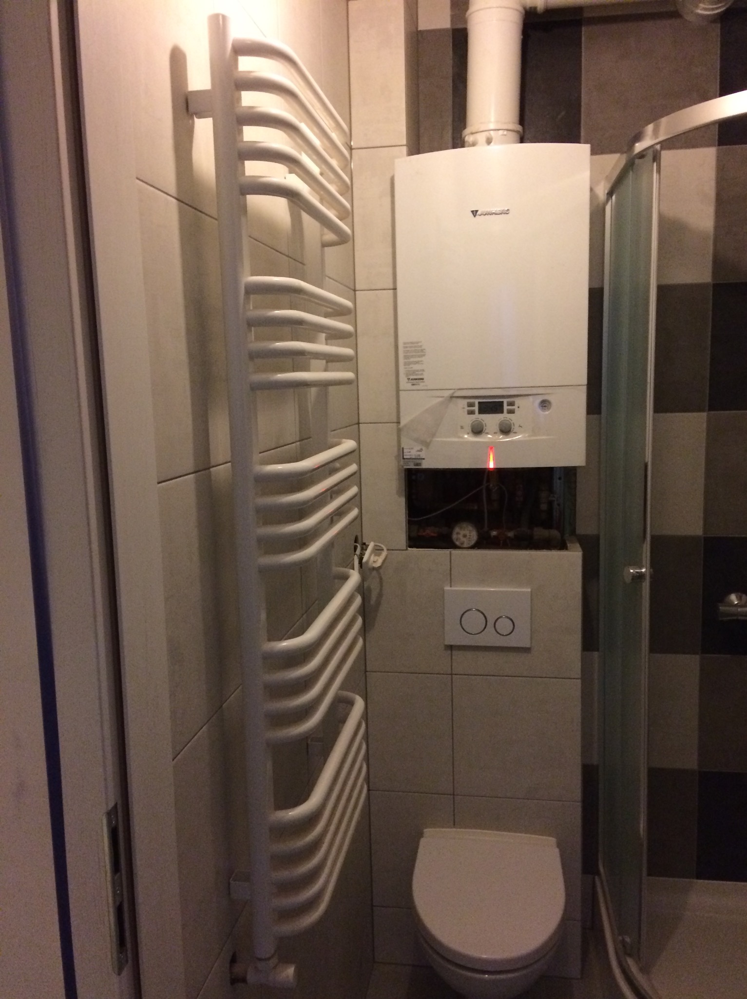 Biała łazienka z grzejnikiem drabinkowym, piecem gazowym i kabiną prysznicową. Widoczne płytki w odcieniach szarości i biała ceramika. Ujęcie z bliska, prezentujące detale wykończenia.