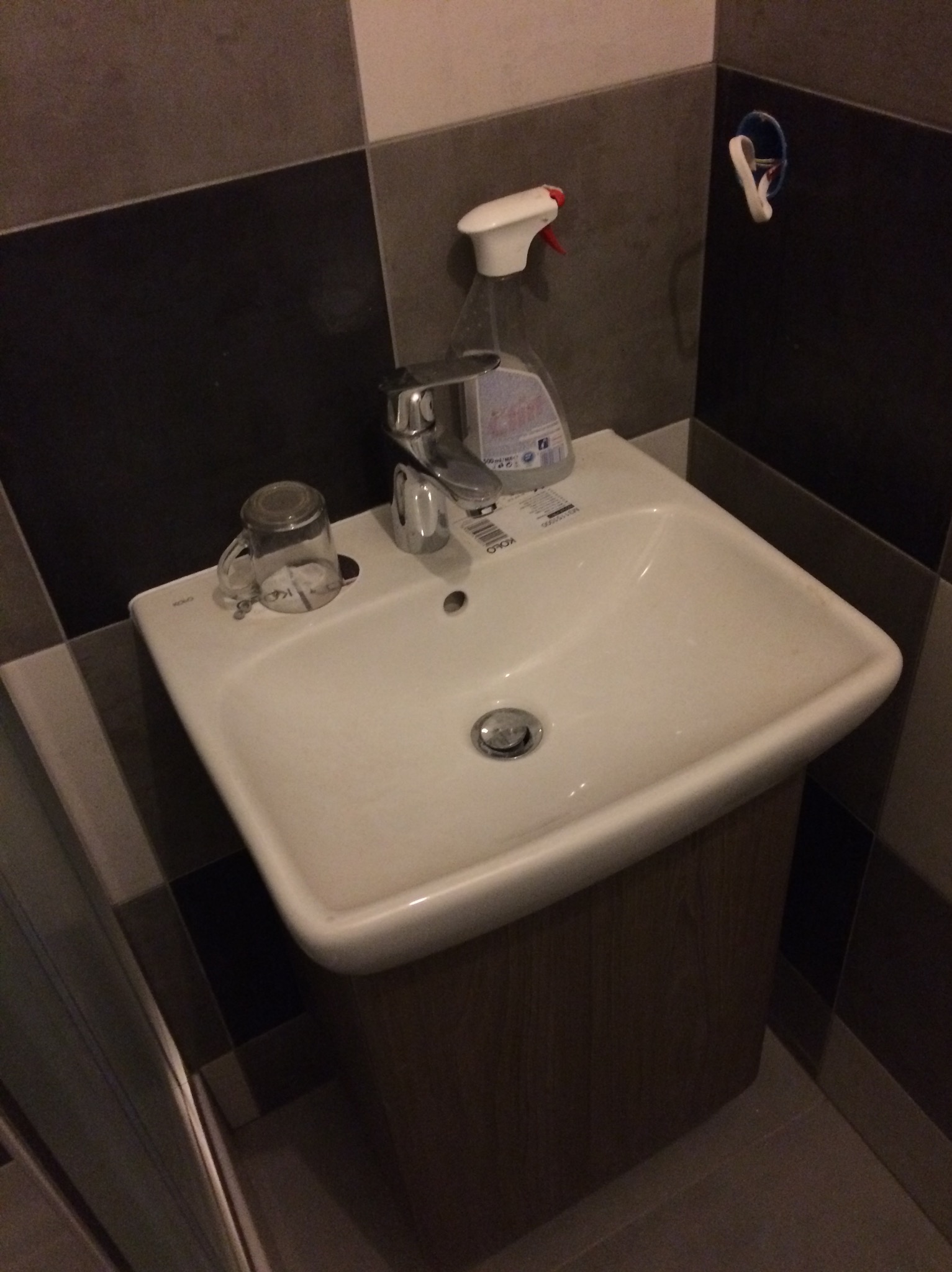 Umywalka nablatowa z szafką w łazience z ciemnymi płytkami. Na umywalce szklanka i płyn do czyszczenia. Widoczne gniazdko elektryczne bez osłony.