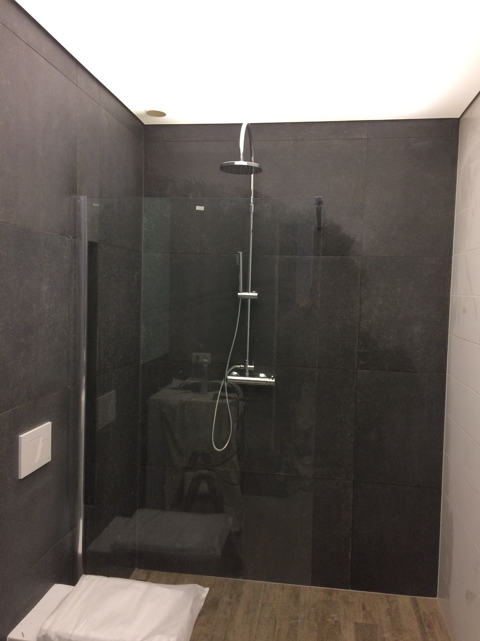 Nowoczesna kabina prysznicowa z ciemnymi płytkami, szklaną ścianką i deszczownicą. Widoczny fragment toalety z poduszką. Minimalistyczny design łazienki.