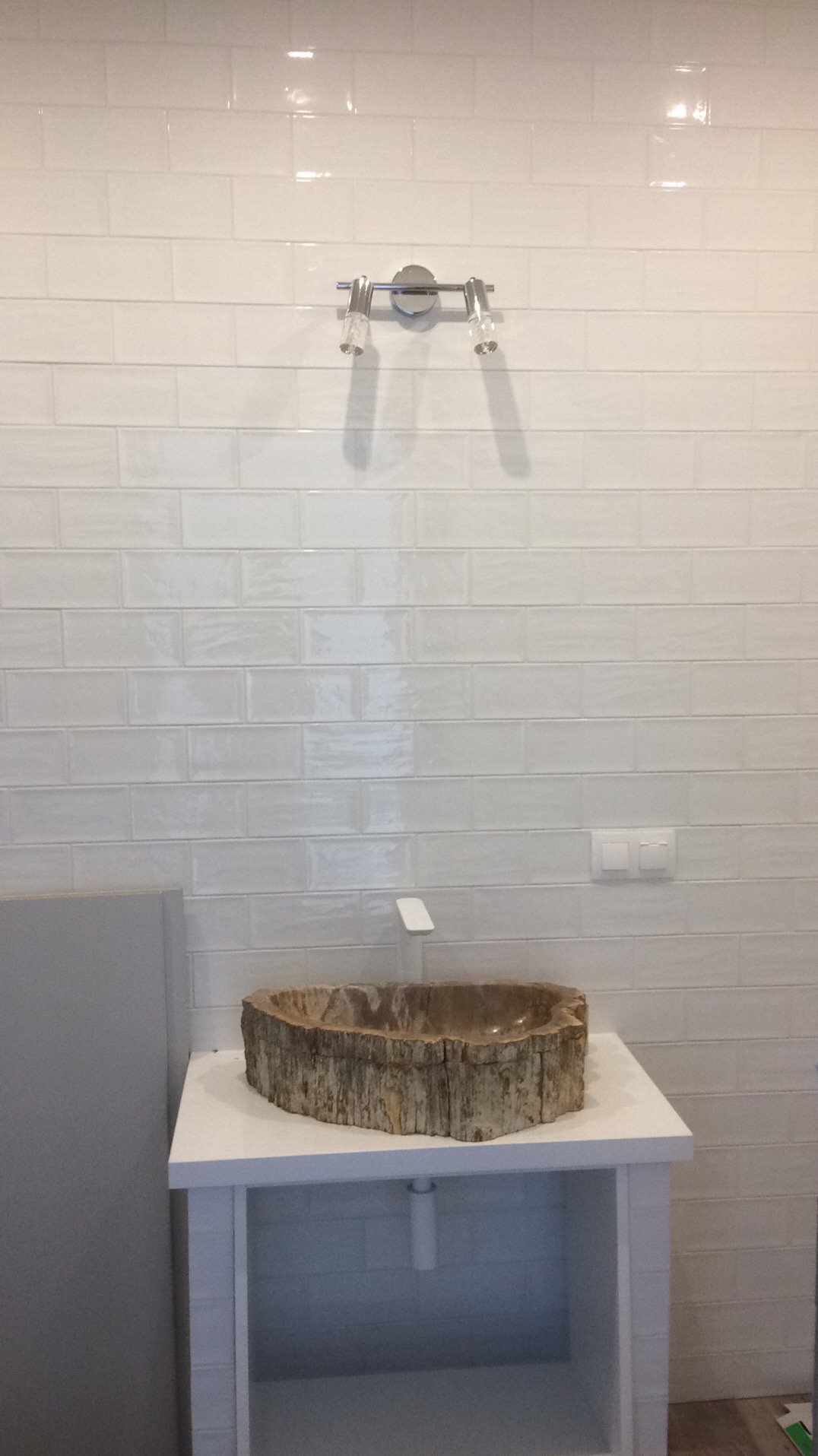 Biała łazienka z umywalką z pnia drzewa na szafce. Na ścianie białe, błyszczące płytki imitujące cegłę i nowoczesne oświetlenie.