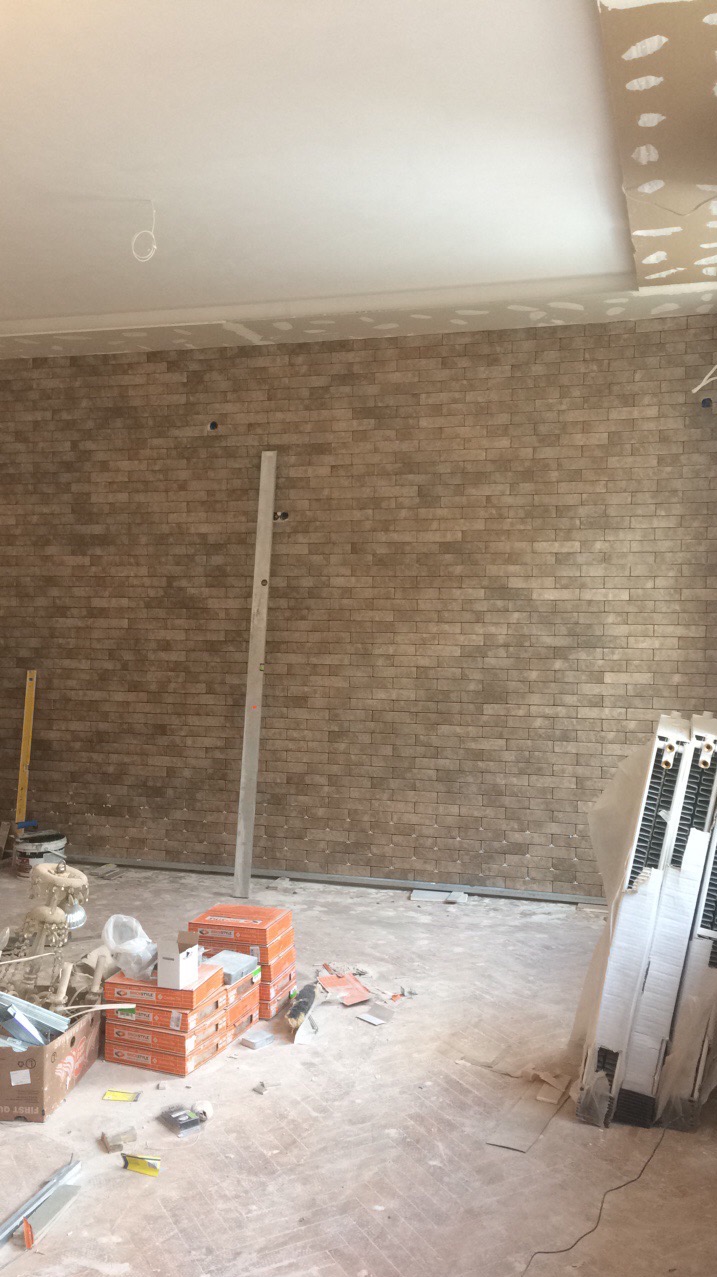 Ściana wyłożona ceglastymi płytkami w trakcie prac wykończeniowych. Widoczne narzędzia, poziomica i materiały budowlane. Sufit w trakcie montażu płyt karton-gips.