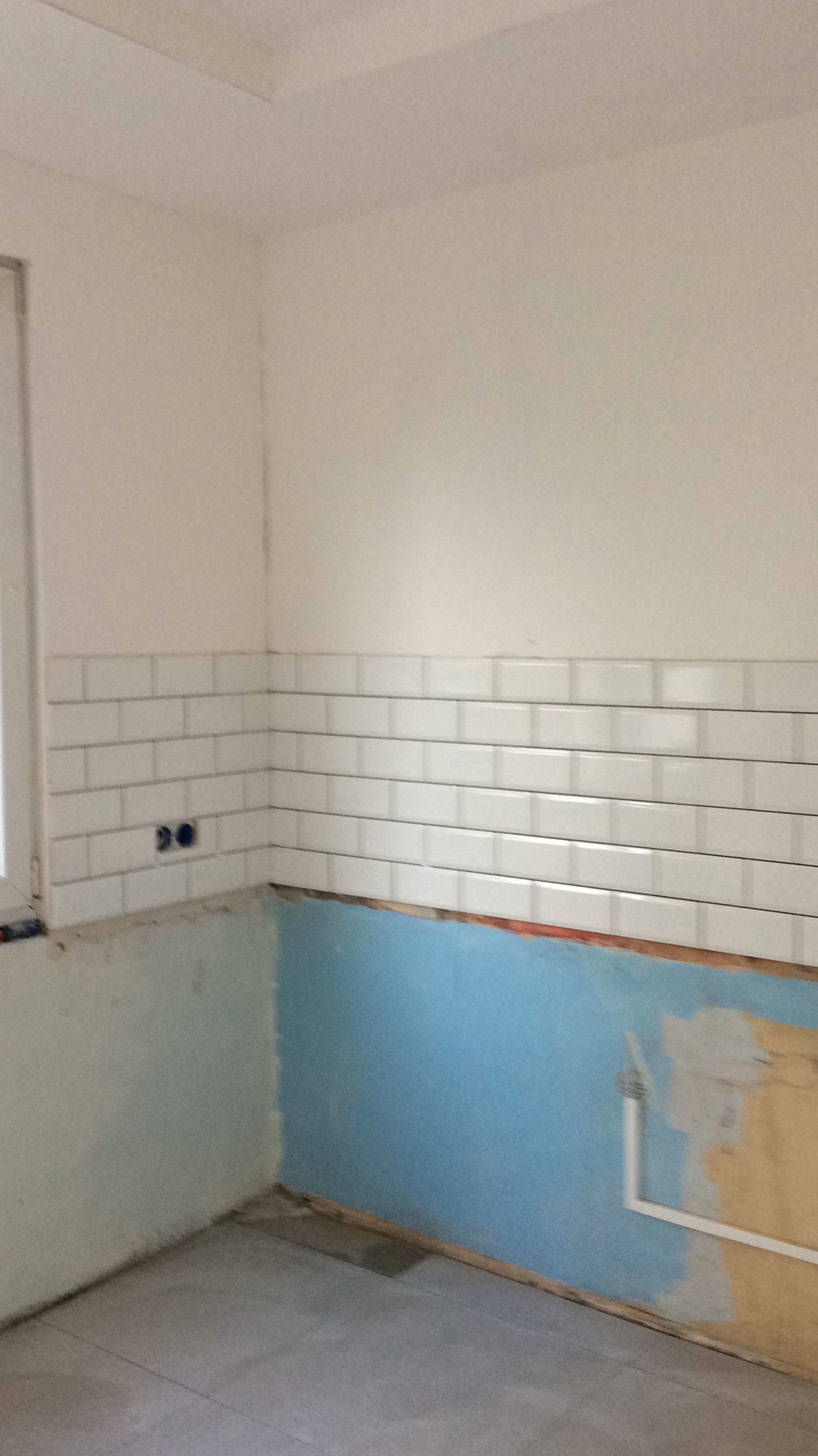 Fragment kuchni w trakcie remontu: białe, prostokątne płytki ułożone na ścianie, niebieska farba poniżej, widoczne gniazdka elektryczne i szare płytki na podłodze.