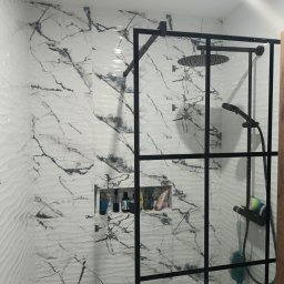 Maiki - Nowoczesna kabina prysznicowa z czarną ramą i deszczownicą, białe płytki imitujące marmur z szarymi żyłami, półka w ścianie z kosmetykami. Wykończenie łazienki.