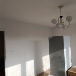 Maiki - Jasne, odświeżone wnętrze z drewnianą podłogą i białymi ścianami, oświetlone przez okno i lampę sufitową. Widoczny fragment lodówki i kartonów.